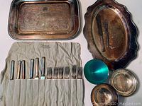 Photo showing two large ornate silver plate trays with detailed engraved patterns, small bowls, and folded cloth holding silverware including knives and forks with leaf design handles.