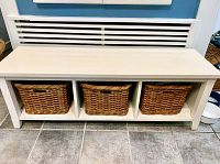 Full view of white wooden bench with three wicker baskets fitted into cubbies beneath the seating surface.