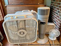 Five fans arranged closely including three large white box fans, a tall tower fan in beige and gray with control panel, and a small white desk fan from Comfort Zone