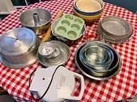 Full lot of baking supplies on a red and white checkered tablecloth including cake pans, muffin pan, pie pans, mixing bowls, rolling pin, and waffle baker.
