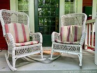 Two white wicker rocking chairs with beige and rust-colored faded striped cushions on a concrete porch