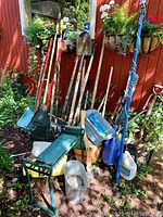 Overall view of various gardening tools and equipment leaning against a shed, including rakes, shovels, pruning shears, a weeding bench, hoses, and containers.
