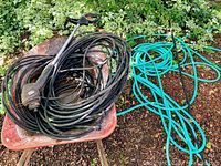 Wide view of metal wheelbarrow with tire, Black & Decker electric trimmer coiled in wheelbarrow plus two turquoise garden hoses and two black soaker hoses on ground beside wheelbarrow.