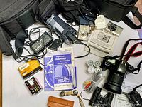 Overview of lot showing Canon cameras, JVC and Sony video cameras, binoculars, accessories, chargers, film rolls, and manuals.