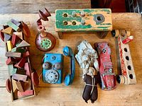 Overview photo of the antique toys lot displayed on a wooden surface. Includes colorful wooden blocks in a truck, two vintage toy phones, a parachutist doll, and wooden pounding bench.