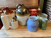 Photo showing all items together arranged on wooden surface with piano in background; the three glazed pottery jugs, blue ceramic pot, green Mason jar, oyster tin can, and two bottles visible.