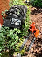 Photo showing large electric blower vac with Black & Decker branding on the leaf collection bag and two blower units lying outside amongst foliage.