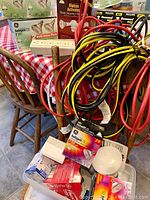 Top view showing a cluster of red, yellow, and black heavy duty extension cords coiled and piled on a wooden chair; boxes of GE halogen bulbs and a motion activation sensor flood light in packaging visible in background on the table; white light globes and other small lighting components arranged below cords.