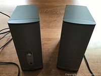 Two Bose desktop speakers on a wooden surface, front view showing metal grille and plastic casing.