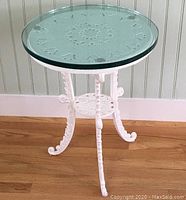 Full view of white cast iron porch table with round glass top showing decorative leg and base design.