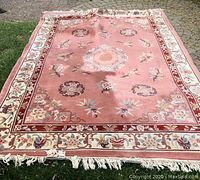 Full view of large rectangular peach/rose colored Chinese rug with floral and medallion design outdoors on greenery.