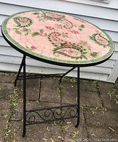 Angled side view of the round patio table showing the ceramic tile top with paisley pattern and black wrought iron foldable base with scroll designs.
