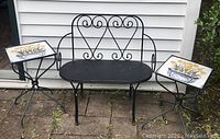 Full view of black wrought iron bench between two matching side tables with floral ceramic tile tops, set outdoors on stone patio.