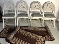 Full view of four white Samsonite folding chairs with padded patterned seats and two rolled Thomasville rug runners laid out on the floor.