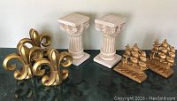 Three sets of bookends on a green marble surface: gold-painted heavy metal sailing ships stamped 'Old Ironsides', white resin Ionic columns, and gold-painted resin fleur de lis.