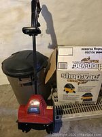 Red Toro Power Shovel with black handle and cord placed in front of a gray plastic trash barrel and Shop Vac box.