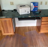 HP LaserJet Pro M203dw printer on a green countertop desk with power cables, two clear floor protectors on wood floor below, and boxes of envelopes on the counter.