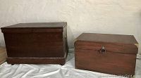 Two solid wooden storage boxes side by side on a white sheet against a wall. The left chest is larger, dark stained, with a plain rectangular design and original hardware. The right chest is smaller, reddish finish, with a front metal ring pull handle and some wear on the edges.