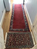 Photo showing long wine-colored runner rug with dark blue floral border and smaller burgundy rug with floral pattern, both with cream fringes.