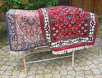 Photo of two Persian rugs draped over the laundry rack outdoors on stone paved area. Shows design, colors and fringe details of both rugs.
