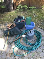 Photo displaying the full collection of gardening items including hose, watering can, buckets, gloves, boots, kneeling pad, and hand tools arranged on a stone surface outdoors.