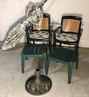 View of 2 forest green patio armchairs with faux caned backs, 2 small green plastic side tables, folded floral canvas umbrella and umbrella base on concrete floor