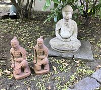 Photo shows the Buddha statue seated on a stone slab with the two terra cotta kneeling archers placed side by side on the dirt ground, surrounded by garden plants and tree branches.