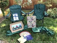 View of two green collapsible nylon outdoor chairs with cup holders, assorted trays including a floral plastic tray, a painted wooden tray with a barn scene, a blue tile trivet, and new plastic tablecloths displayed on the grass.