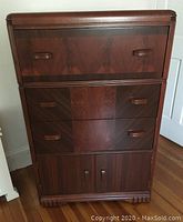 Front view of the Art Deco dresser showing three drawers with wood veneer faces and two-door cabinet at bottom, wood pull handles on drawers and doors, standing on wooden floor against wall.