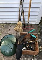 Photo showing a vintage wooden box filled with various small gardening tools, a broom with natural straw bristles, a metal rake with wooden handle, and green garden hose head, all on stone pavers against white siding.