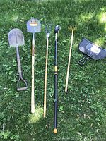 Full view of all six lawn tools laid out on grass showing shovel, toothed shovel, rakes, hoe, and dustpan.
