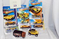 Seven Hot Wheels cars mint on card displayed with two open stock vehicles in front, all in original packaging or loose condition, shown against a white fabric backdrop.