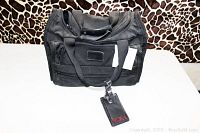 Full front view of the black TUMI weekender bag showing multiple zippered compartments and attached leather luggage tag.