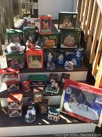 Photo showing wide view of many boxed and unboxed porcelain lighted Christmas village houses and accessories, including animated skating pond box on right bottom.