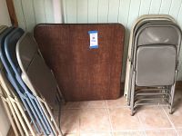 Photo of a folded rectangular vinyl top folding table leaning against the wall between stacks of folding chairs in beige and gray.