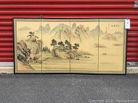 Full view of four panel folding screen with traditional landscape painting and decorative border, set against red storage unit backdrop.