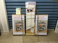 The full lot showing two boxed 3-tier wire shelving units, the gold metal free-standing towel rack with three hanging bars, the fabric wardrobe box behind the towel rack, and the Marie Kondo book.