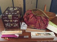 Front view of wooden jewelry box with mother of pearl inlay, red silk purse with gold embroidery, two boxed wooden folding fans, and silk scarf laid out on table.