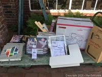 Overview of lot on outdoor bench showing machines in boxes, thread organizer, hoops, spray adhesive, Magic Box kit, printed patterns, magazines, extension table