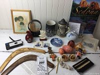 Full view of lot showing all items arranged on table including framed art, boomerangs, paper cuts, stein, eggs, figurines and camera