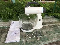 Sunbeam Mixmaster stand mixer with glass mixing bowl, accessories, and instruction manuals on outdoor bench.