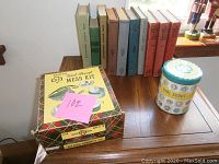 Photo of nine vintage Girl Scout books arranged in a line behind a boxed Girl Scout metal mess kit on a wooden table.