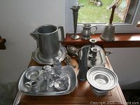 Full view of pewter serveware including a large pitcher, a bell with wooden handle, candlesticks, and a tray with additional smaller items.