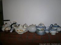Wide view of over 10 ceramic teapots arranged on brick mantle including floral patterns, white and blue colors, some with matching cups and plates
