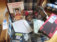 Wide shot displaying Elvis Presley cookbook, trivia game, pin badge, DVD set, and other memorabilia on a plaid couch