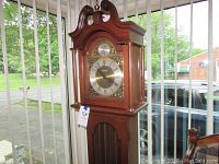 Full view of wooden grandmother clock near windows, showcasing overall shape, finish, and carved details on top.