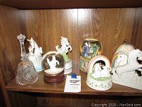 Seven unicorn figurines, two snow globes, and a clear etched glass bell displayed on a wood shelf.