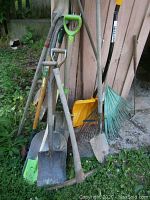 Full view of a collection of shovels and rakes leaning against a wall outdoors, showing condition and variety of tools.