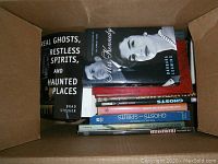 Box containing multiple books related to ghosts and strange phenomena. Titles visible include 'Real Ghosts, Restless Spirits, and Haunted Places', and a biography of Rose Kennedy.
