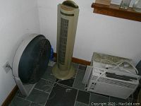 Three cooling appliances placed on a tiled floor against two walls: a black circular fan on gray base, beige tower fan, and a white window air conditioner.